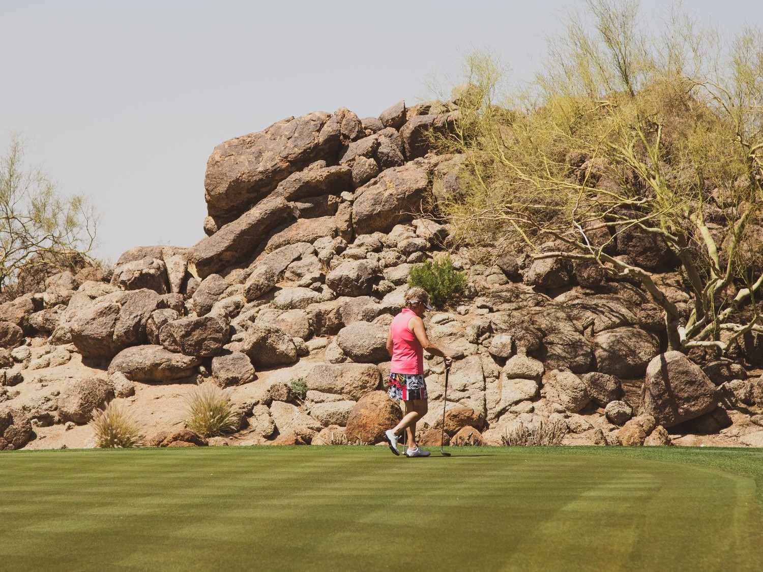 A golfer on a green fairway in front of a rocky desert hillside with palo verde trees