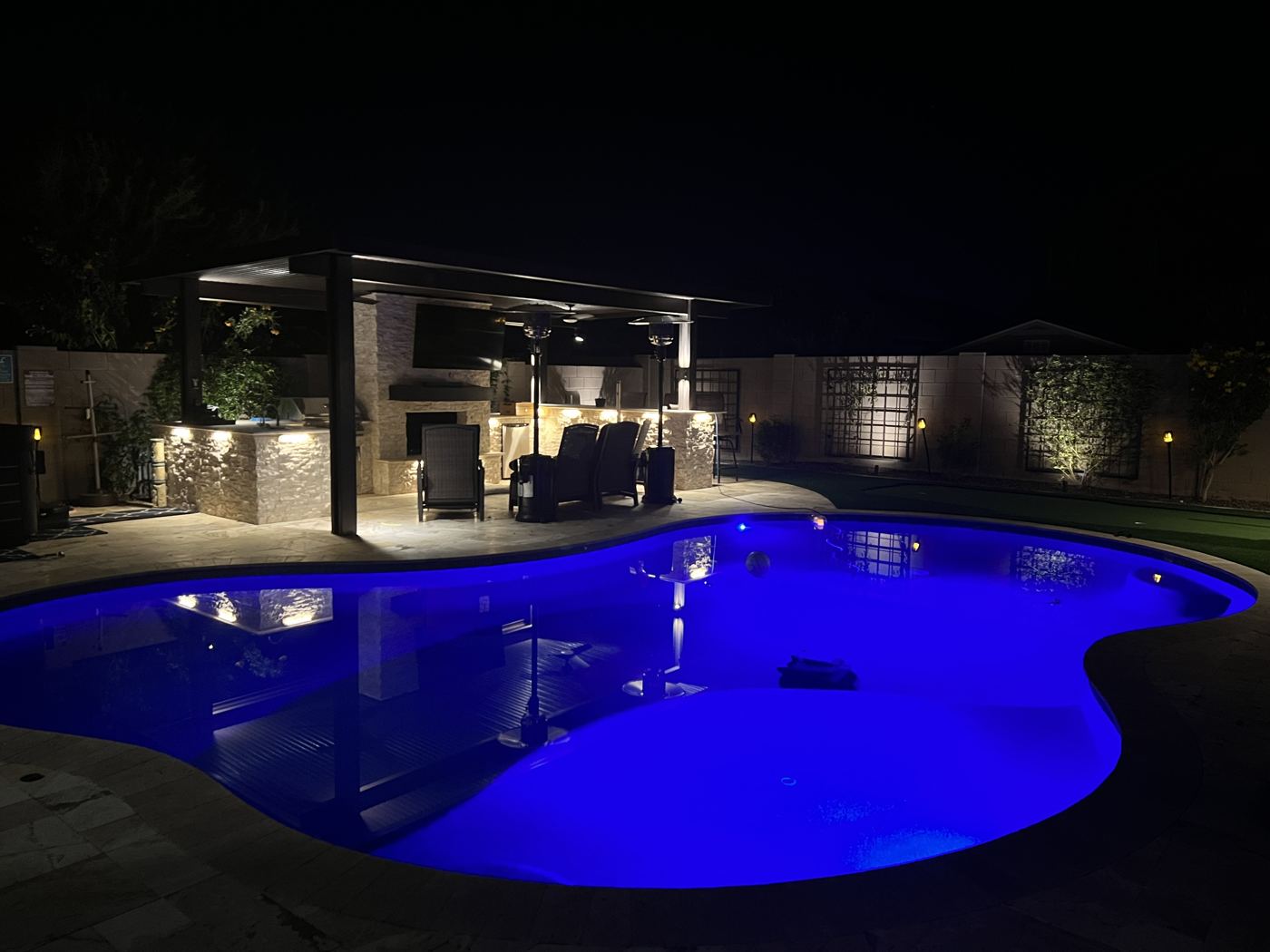 A backyard pool lit blue at night beside a covered outdoor kitchen